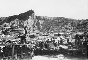 A view of the beach north of Anzac Cove. In the foreground some men are standing on Williams' Pier at North Beach, and a crowd of others is nearby. Between the pier and a wharf is a K lighter, used to transport stores. There are some tents and many dugouts on the slopes rising behind the beach. Dominating the scene at left is the rock formation called The Sphinx.