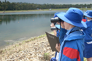 A group of young students standing on a bank and looking through binoculars across water