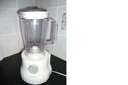 A blender with a white hard plastic base and grey dial. A clear plastic bowl with a handle and a white lid is attached to the base