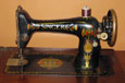 A black metal sewing machine decorated with a red and gold design; it has  brown cotton on the spindle.