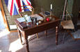 A teacher's desk covered with classroom items with a chair and part of a flag in the background.