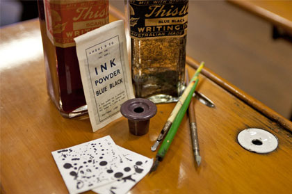 Two bottles of ink and instructions for mixing ink powder, brown ink well and a white one set in the desk top. Three pens with metal nibs and two squares of blotting paper.