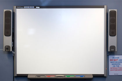 An interactive whiteboard with a speaker on each side. A shelf below the board has a black, red, green and blue marker pen with a whiteboard eraser.