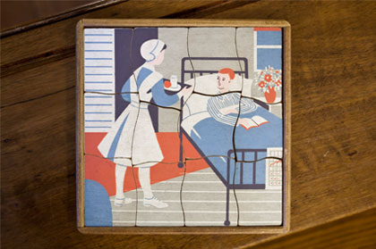A 12-piece wooden jigsaw puzzle showing a nurse in a blue uniform, white apron and cap bringing a tray with fruit and a drink to a patient in hospital. 