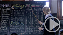 Teacher in early days clothing using a cane to point to writing on a chalkboard