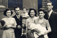 A group of five adults standing in the street; a woman is nursing a baby while a man is holding a toddler.