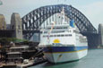 A large white cruise ship with a blue stripe berthed at the overseas terminal Sydney with the Harbour Bridge in the background.
