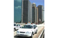 A moving white taxi with tall buildings in the background.