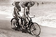 Two women on a tandem bike wearing bathers and holding umbrellas.