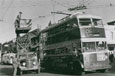 A double decker bus; there are workmen on an old cherry picker fixing the electric wires.