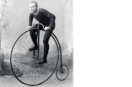 A penny farthing bicycle with a very large front wheel and a small back wheel. The handlebars are above the large wheel.