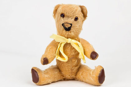 An old teddy bear with orange fur, brown glass eyes, four paws and a yellow ribbon