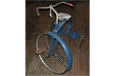 A rusty blue metal tricycle with white seat and red hand grip.