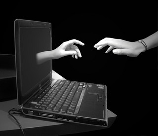 Black and white image of a hand reaching from a laptop screen towards someone else's hand