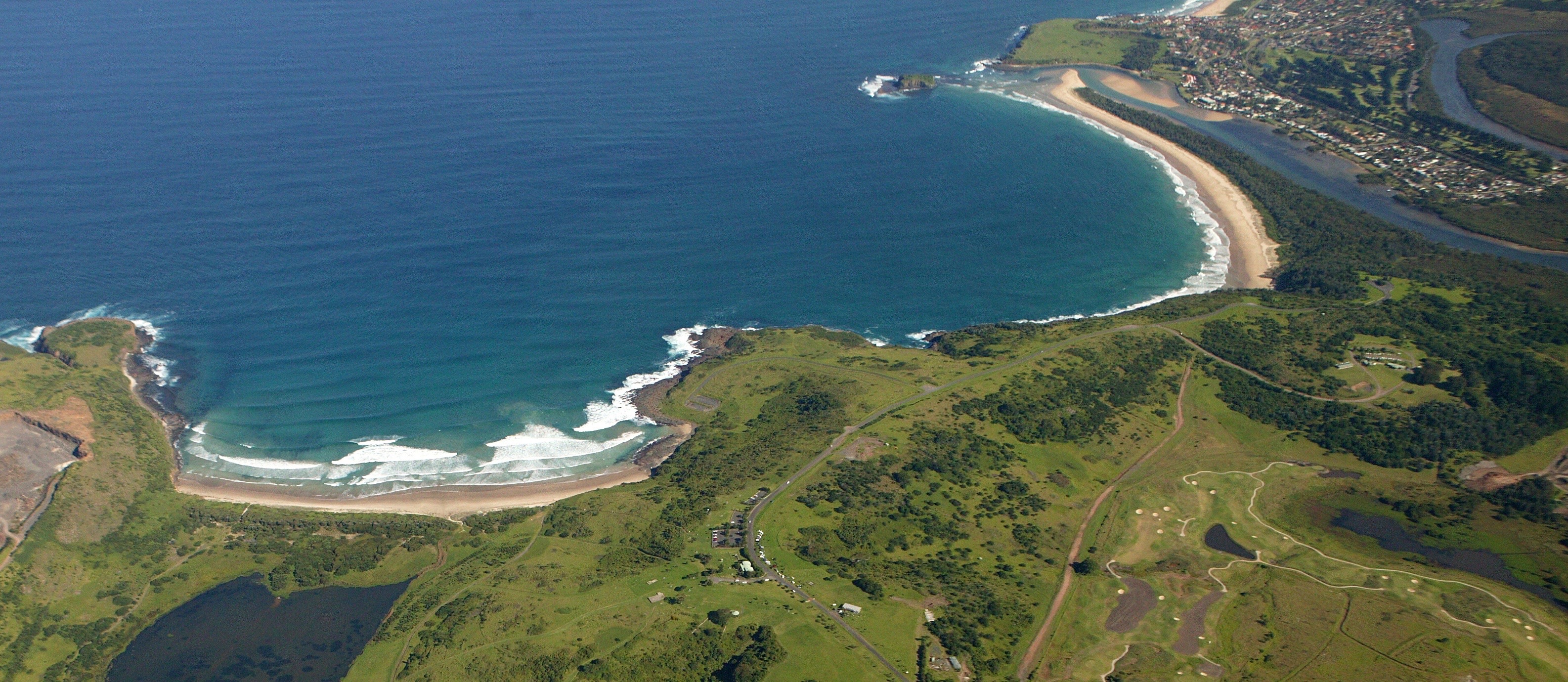 Aerial view of the coastline with deep blue sea, white sandy beaches, some rocky areas and then grassy land behind the beach.