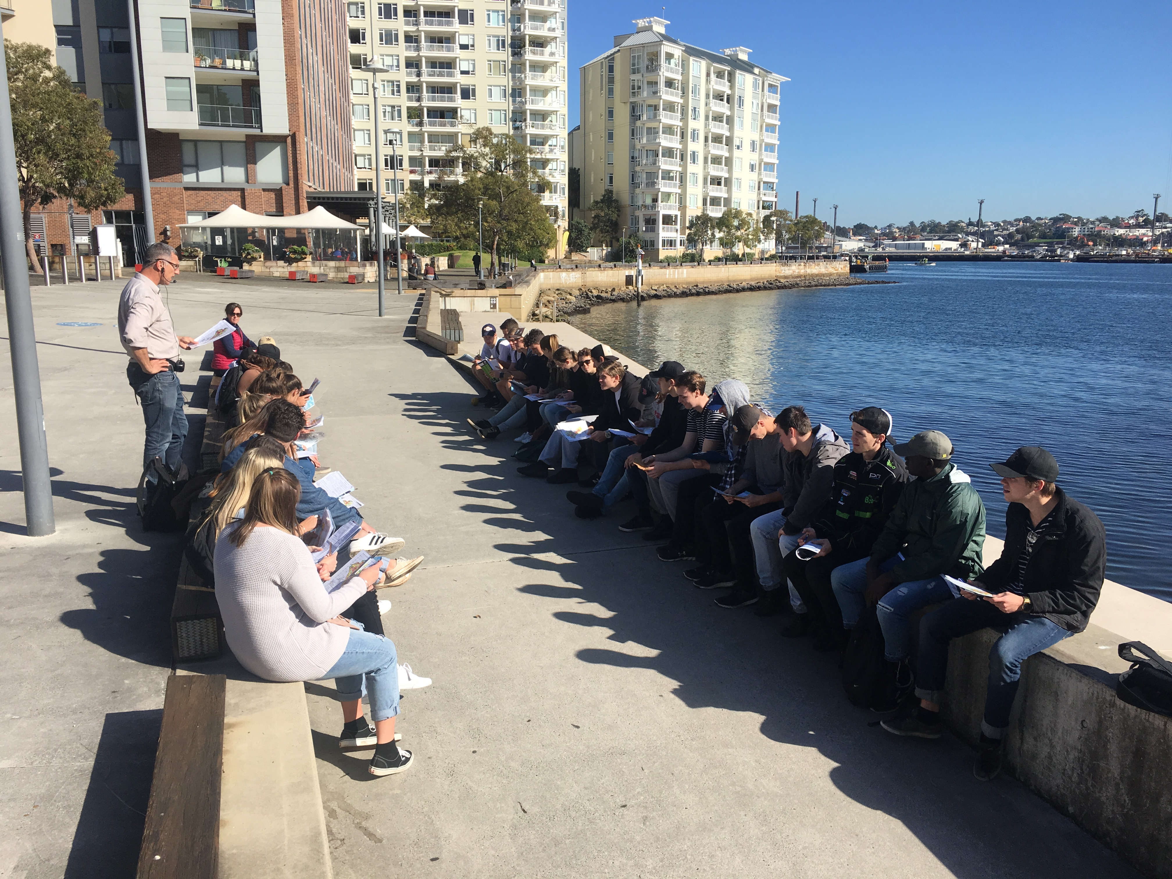 large group opf stduent sitting on a concrete wall built on the foreshore of syndey harbour. They are listening to a teacher.