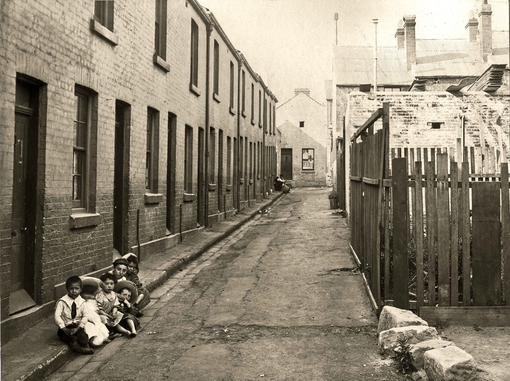Black and white photo showing a narrow side lane with a cabinetmaker's workshop on one side and several poor-looking residential buildings on the other. Five children sit on the curb on the left-hand side of the photo.