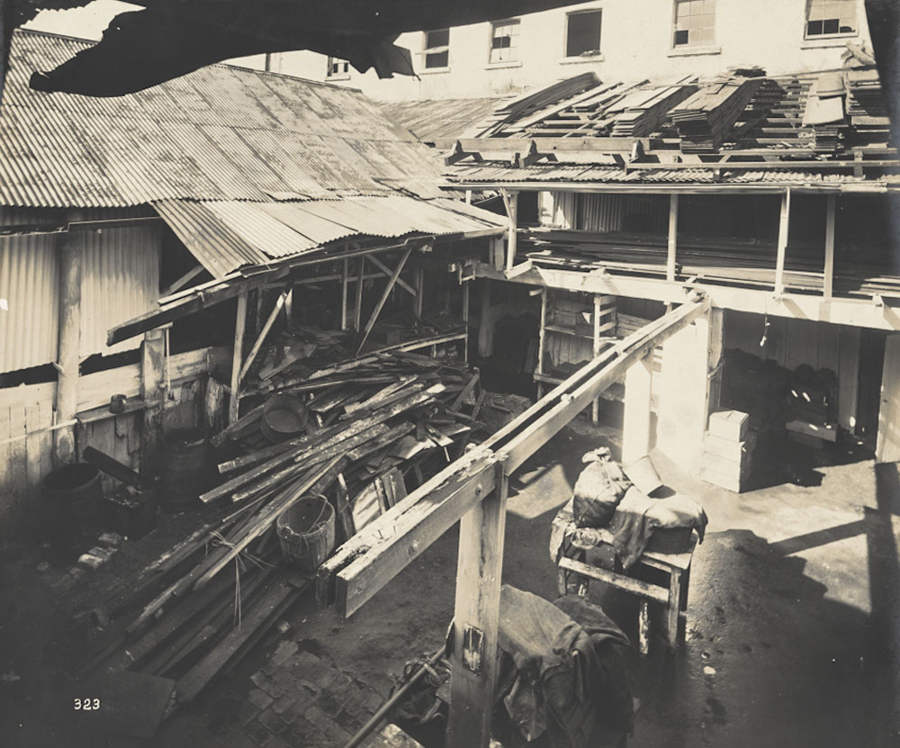 Black-and-white photo showing cabinetmaker's workshop and yard. The yard and roof are cluttered with wood planks as well as a table, several buckets and worker's tools.