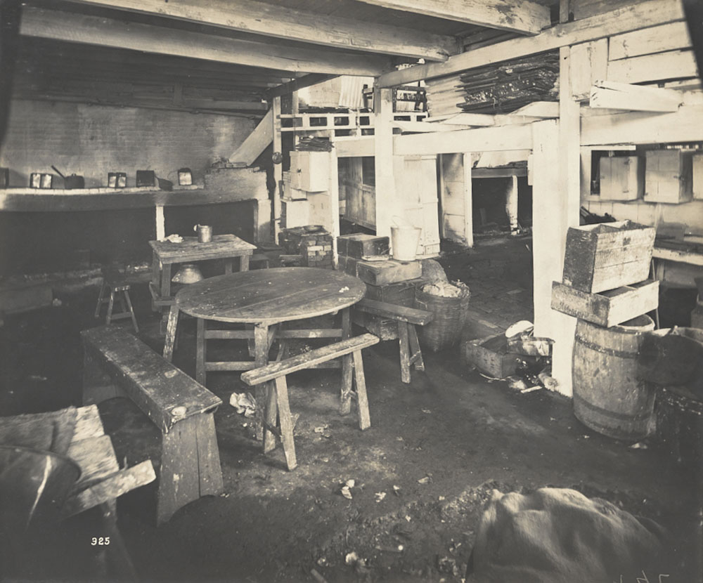 Black and white photo showing a cabinetmaker's workshop. The rooms have concrete floors with some rocks scattered around and includes tables, chairs, some boxes and barrels.