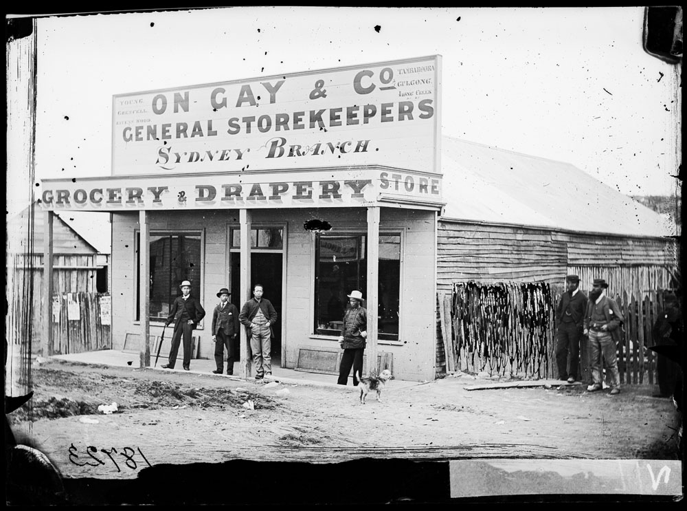 Photo of grocery and drapery store. Signage on top says, 'On Gay and company general storekeepers Sydney branch'. Four men stand in front of the store and two on the right hand side.