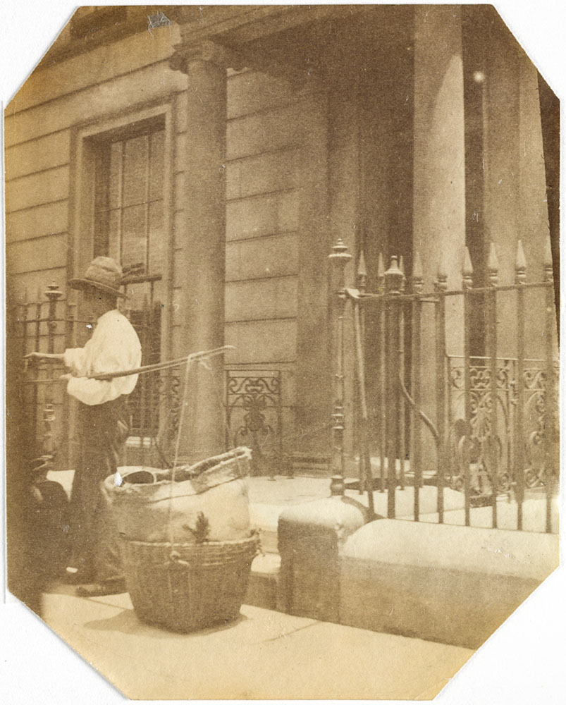 Photo shows a Chinese hawker standing in front of a house with his basket of goods.
