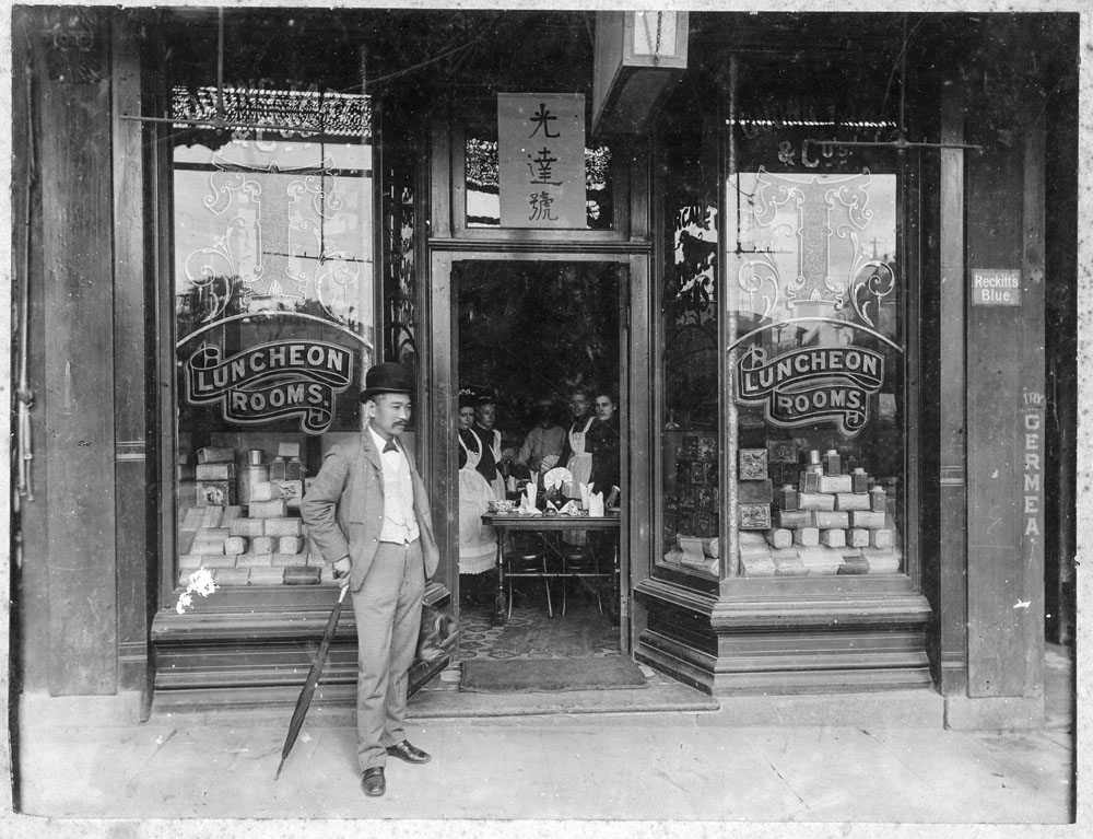 Photo of Quong Tart standing outside one of his tea rooms. Inside are several employees standing around a table.