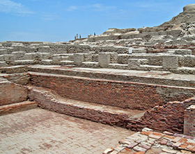 Mohenjodaro archaeological site with sunken bath in the foreground and brick structures surrounding it and mound in the right background.