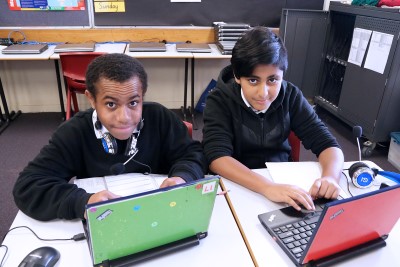 Decorative image of 2 children in a classroom with laptop computers.