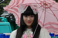 Fashion thumbnail: A woman with dark hair and large bow on top, and wearing a white dress; she is carrying a frilly, pink umbrella as she smile at the camera