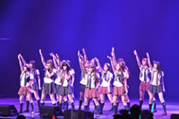 J-pop thumbnail: about 15 girls stand on stage holding microphones and with one arm pointing skyward