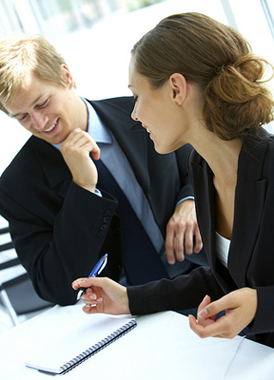 A man and a woman dressed in suits in an office environment. They are talking. The woman has a notebook in front of her and a pen in her hand ready to write.