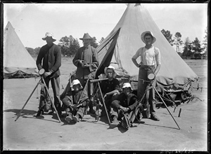 Six men standing or sitting in front of tents