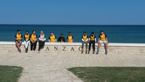 Ten students sitting on a low wall with the word ANZAC on it. The sea is behind them.