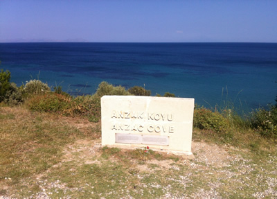 A plaque ANZAK KOYU/ANZAC COVE— on a grassy cliff top overlooking water.