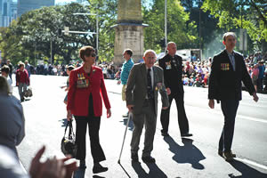 An elderly man walks with the aid of a walking stick down a Sydney street lined with people; he is flanked by a woman on one side and two men on the other; they are all wearing medals