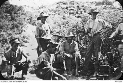 A black and white photograph of 8 men, some sitting, some standing, some eating, some drinking.