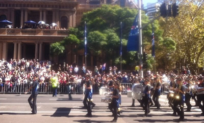 A band marching down George Street past the Sydney Town Hall. Many people line the street, and steps and balcony of the town hall.