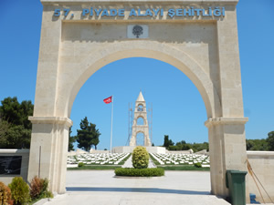 In the foreground is a large archway with the Turkish words, 57. Piyade Alayı Şehitliği (Cemetery of the 57th Turkish Regiment). Behind it are rows of grave stones set in a lawn and a large monument and a flagpole flying a Turkish flag.