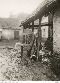 A soldier sits one the edge of a verandah, surrounded by muddy ground, writing on note paper