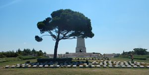 Cemetery plaques on a lawn with a pine tree and momument in the background.