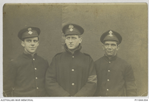 Three men in military coats and caps standing in front of a bare wall.