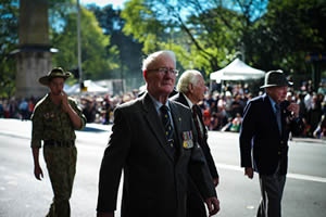 Three elderly men march down the street. You can see that one is wearing medals. They are followed by a youth wearing an army uniform. People line the street.