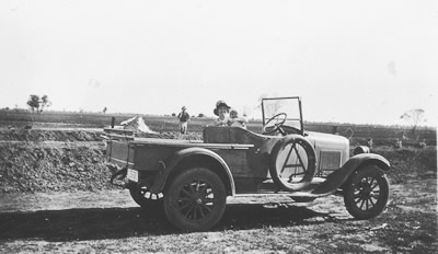 A woman holding a baby in the front seat of a vintage truck in front of the a wide open paddock. A man is standing in the wide open paddock behind them.