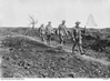 Five men walk along a barren path, the first holds a white flag, the next two carry a man on a stretcher.