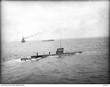 Two men stand on the deck of a submarine; two other naval vessels are in the distance.
