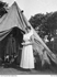 A nurse in a white apron stands in front of an open tent; some clothes are hanging inside.
