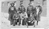 Seven soldiers, spattered with snow, pose in front of a tin barracks; centre front is an Indigenous serviceman