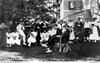 A group of men and women, some seated some standing, drinking tea in a backyard garden.