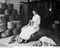A woman sits on a chair knitting. A fleece is beside her and bales of socks are stacked on the left.
