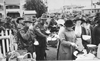 Soldiers stand around eating, holding tea cups, while women are serving out food.